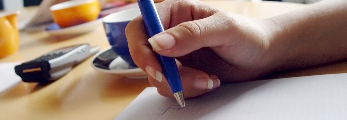 A hand holding a blue pen writing on a paper, with a blue teacup in the background, showing how an organization should also be reviewing its strategic plan.