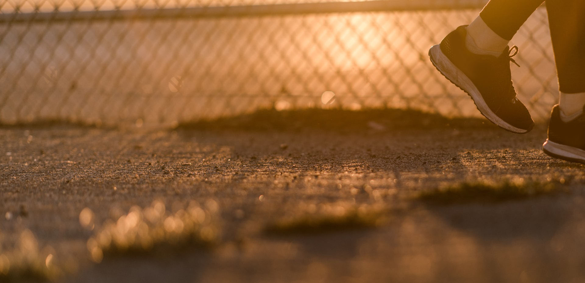 A sepia-toned image of someone wearing sneakers as they walk beside a chain link fence.