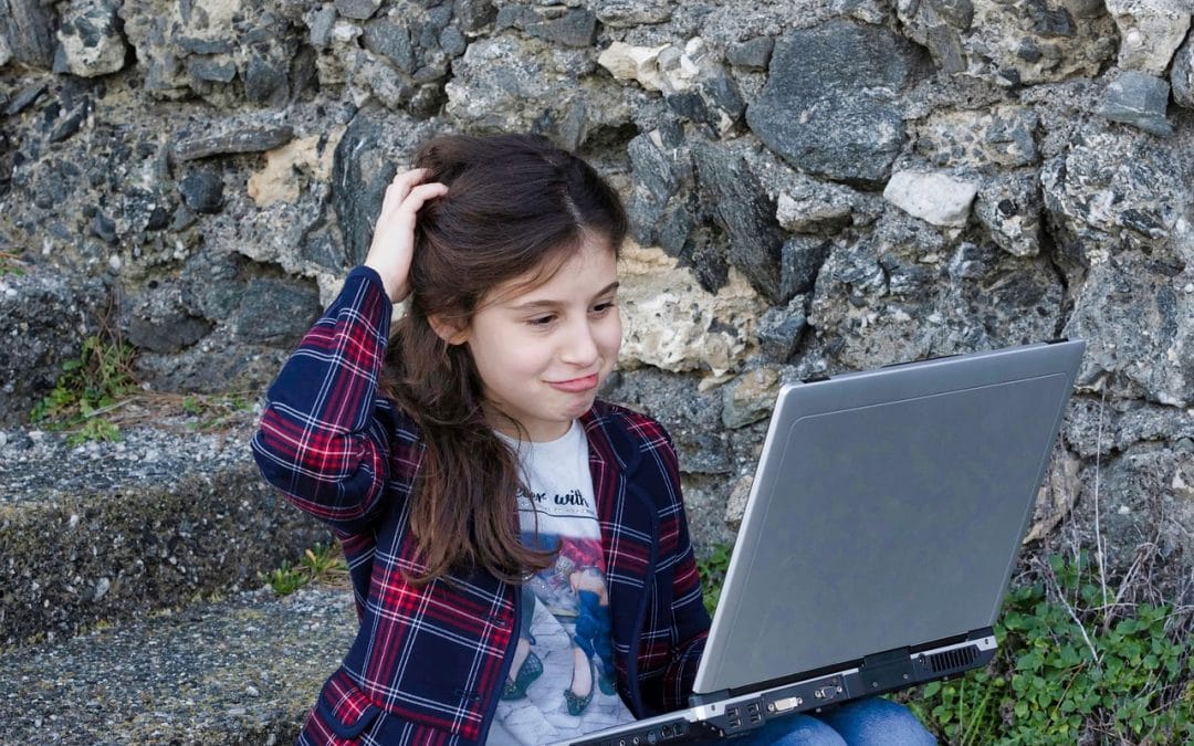 A girl in a plaid shirt sits against a stone wall, scratching her head as she looks at a laptop screen.