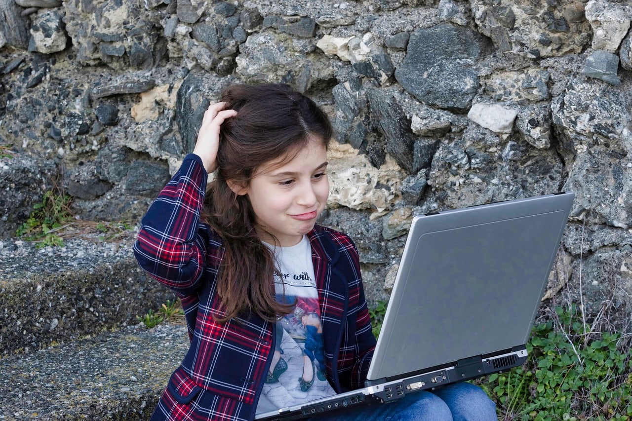 A girl in a plaid shirt sits against a stone wall, scratching her head as she looks at a laptop screen.