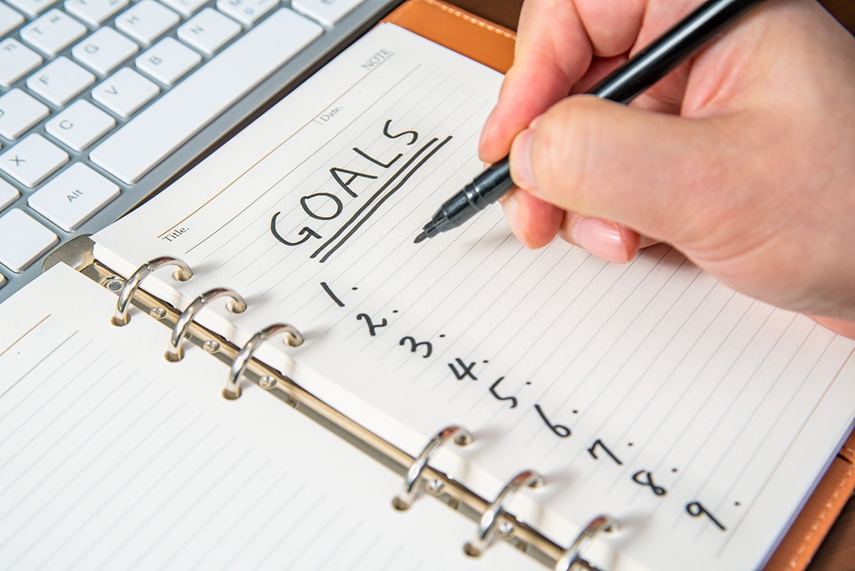 A photograph of a hand writing a list of goals in a notebook.