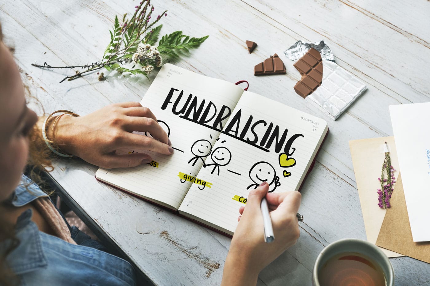 An image of an open notebook on a wooden table, with hands drawing an image of two stick people under the word fundraising, illustrating how a strategic plan can be used to boost fundraising.